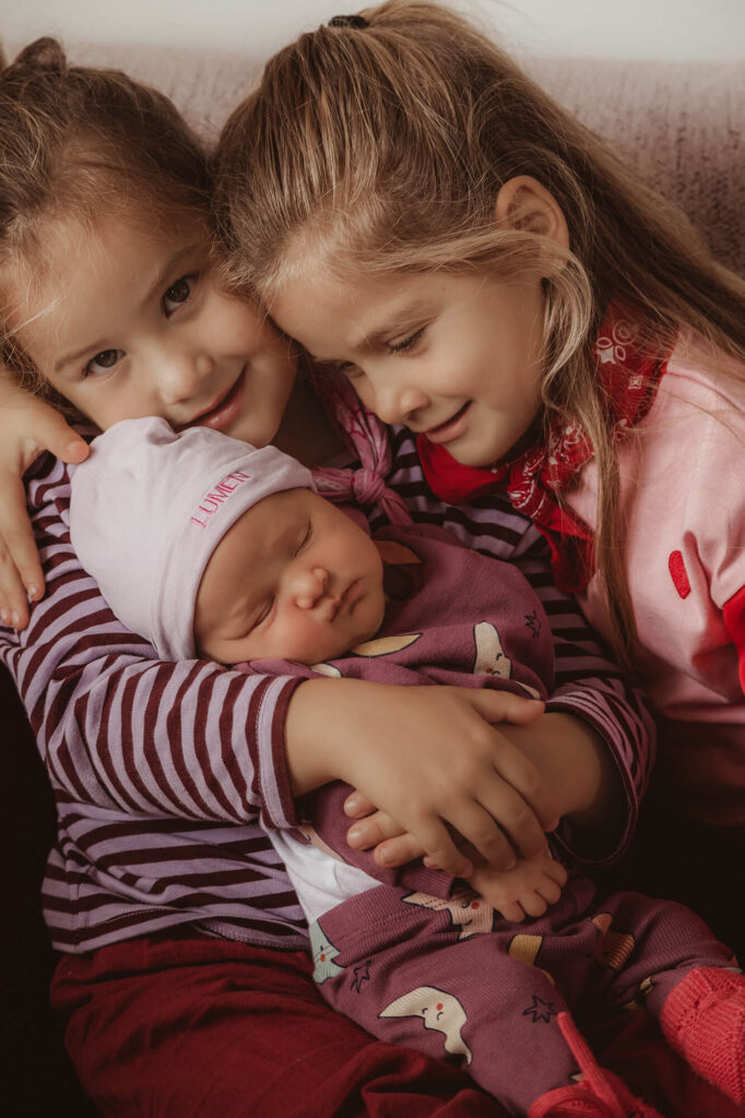 Twee grote zusjes houden de slapende baby vast en kijken lachend in de camera