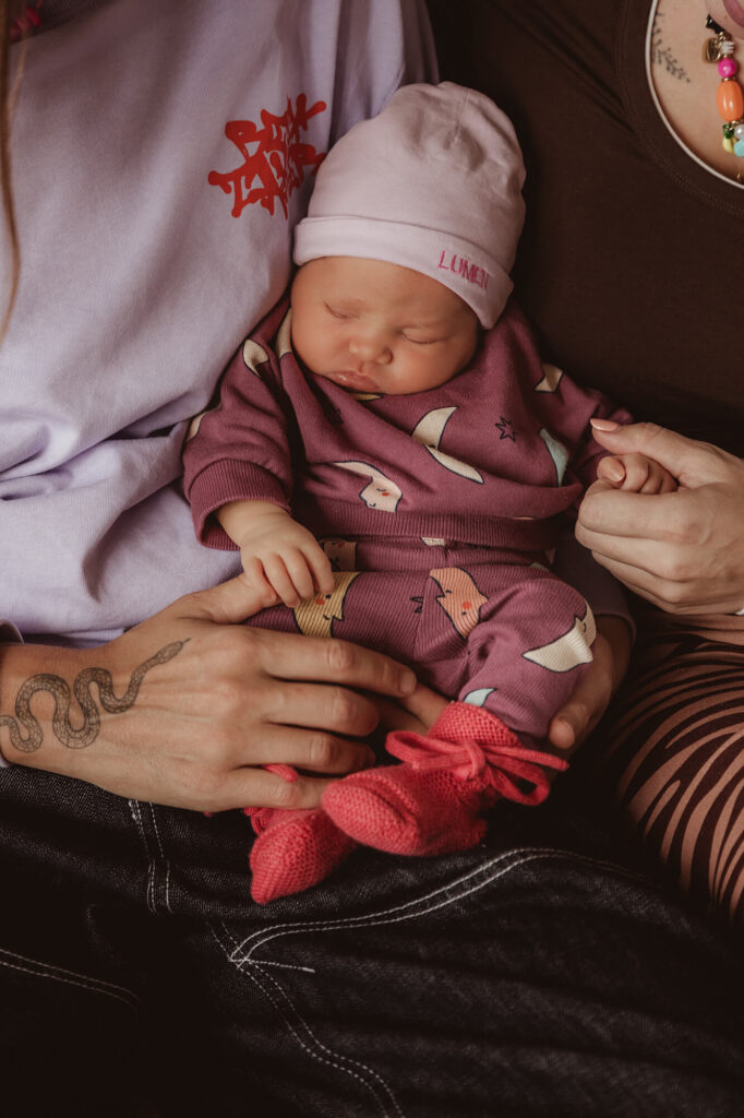 Pasgeboren baby in een lila mutsje houdt een vinger vast bij de ouders op schoot