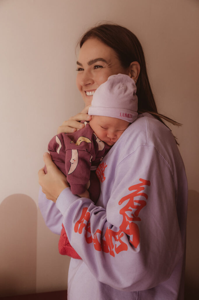 Newbornshoot thuis voorbereiden: lachende mama met haar slapende baby in een lila mutsje tegen haar schouder