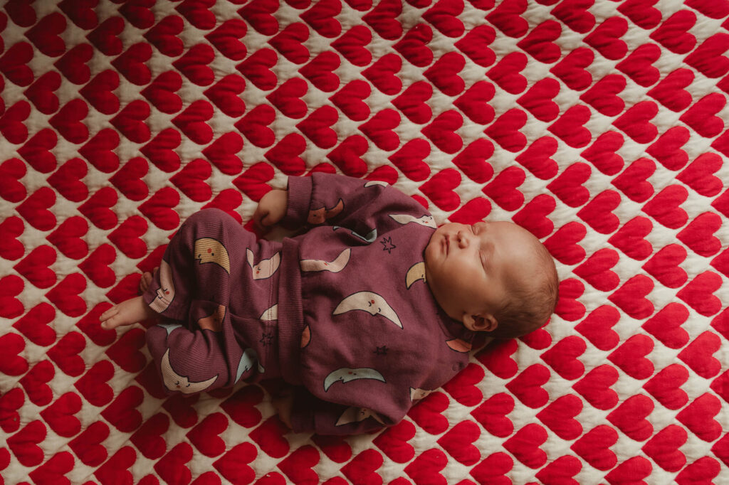 Newbornshoot thuis voorbereiden: baby in een paars maantjespakje slaapt op een quilt met rode hartjes